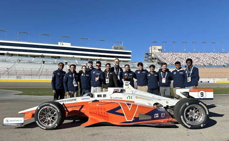 13 people stand beside a race car in a large empty racing stadium
