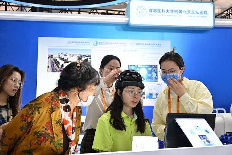 A young woman in a green shirt sits with a wired contraption on her head as four other people look on.