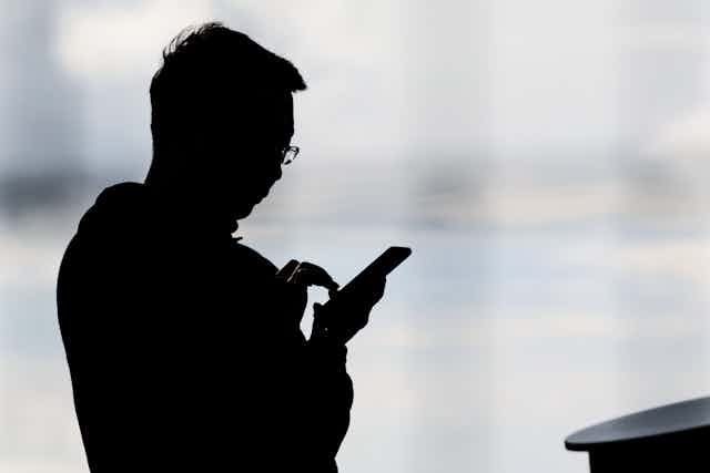 Person with glasses silhouetted against a window using a smartphone
