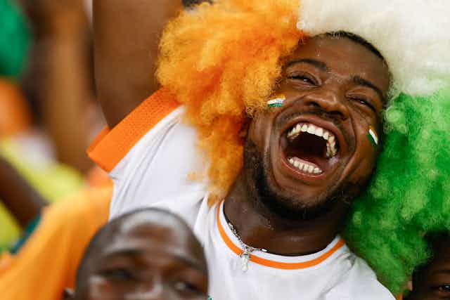 A man's face adorned in a wig of orange, white and green. His mouth is open in a huge smile, his eyes wrinkled with glee.