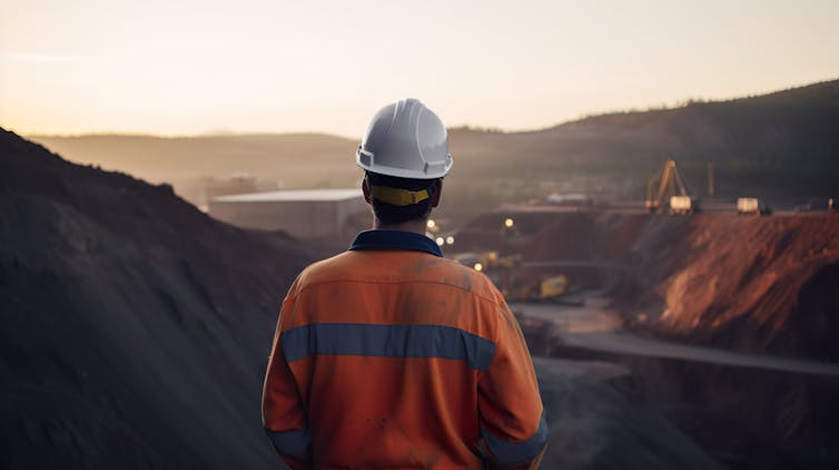 Back of man in orange hi-vis jacket and white hard hard looking across to copper mine, brown roads and pits, grey sky