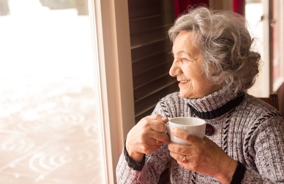 An elderly woman smiles while looking out the window and enjoying a cup of coffee or tea.