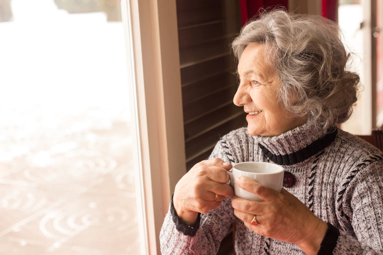 An elderly woman smiles while looking out the window and enjoying a cup of coffee or tea.
