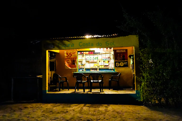 A small building with a verandah and a window to a room with shelves, where a light is shining. Outside it's dark.