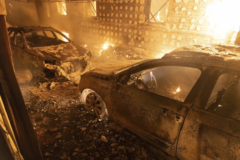 Burnt out cars are seen in front of a flaming structure.