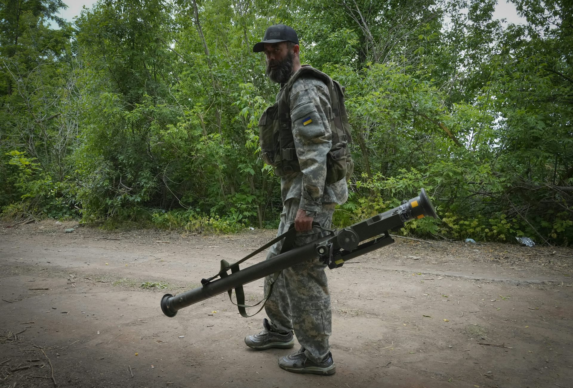 A bearded soldier wearing camouflage carries a large weapon as he walks along a dirt 