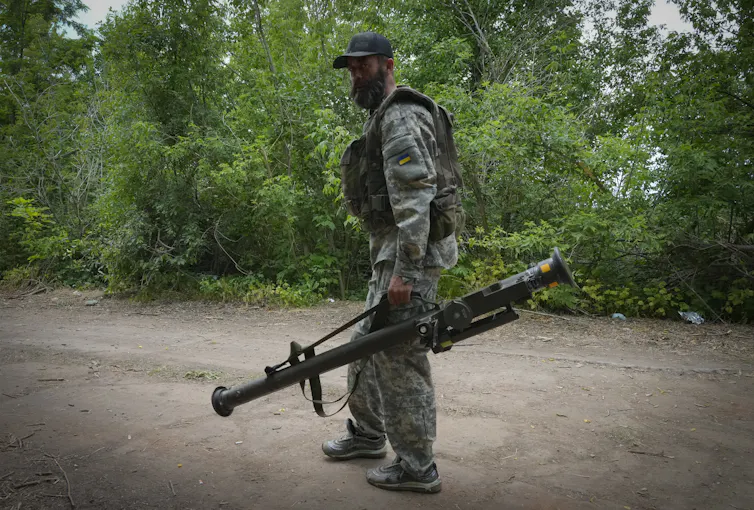 A bearded soldier wearing camouflage carries a large weapon as he walks along a dirt