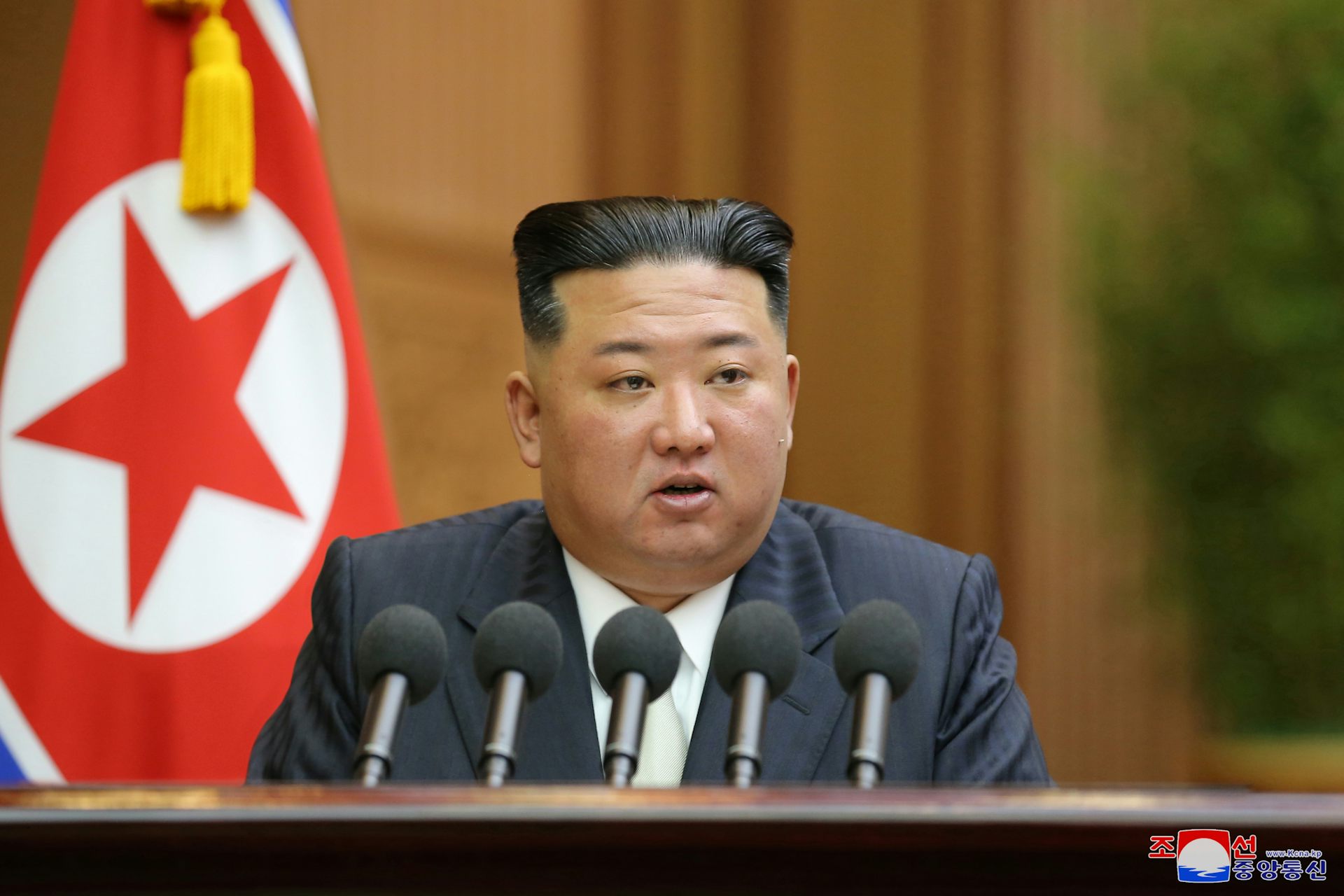 Kim Jong-un sat at a desk delivering a speech in front of the flag of North Korea.
