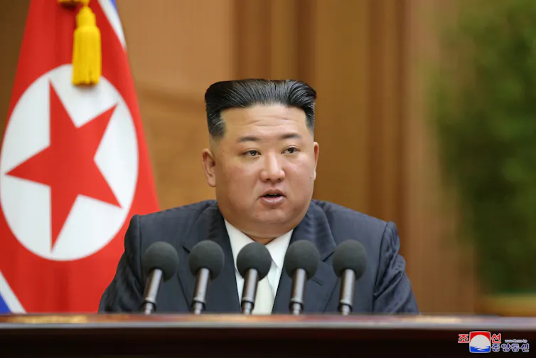 Kim Jong-un sat at a desk delivering a speech in front of the flag of North Korea.
