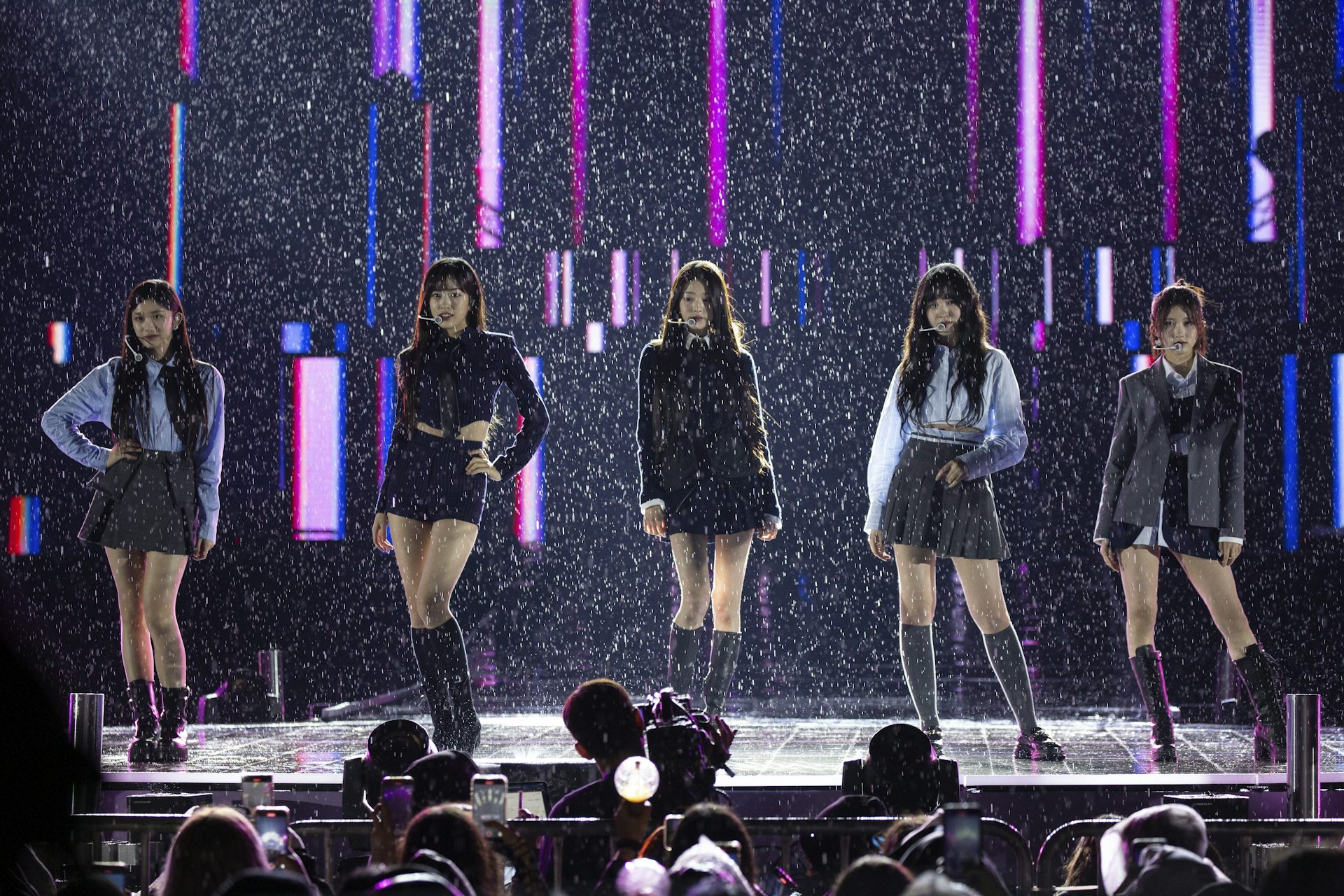Five members of a K-pop girls group standing on a stage performing.