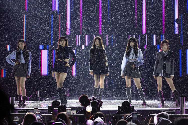 Five members of a K-pop girls group standing on a stage performing.