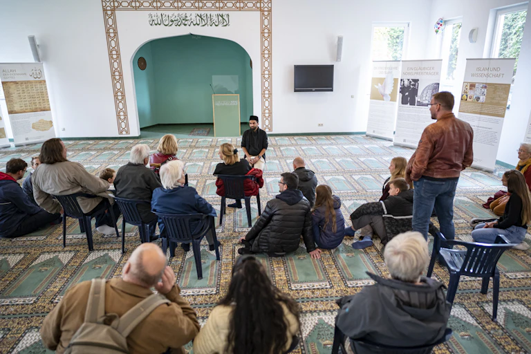 A man in a black jacket and cap talks to people seated on chairs in front of him. Behind him is a wall with a green-color niche, with inscription in Arabic above it.