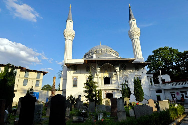 An ivory-colored German mosque with two tall minarets with a dome in their center, set against the backdrop of a clear, blue sky. 
