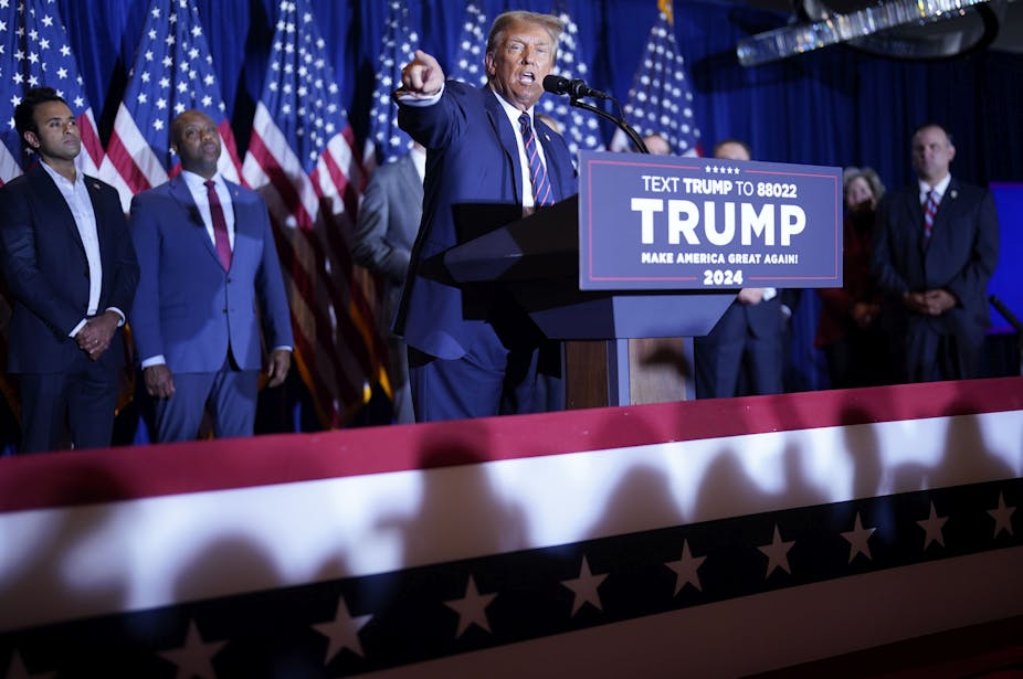 Donald Trump speaks at a podium that says his name, standing in front of a row of people, including Vivek Ramaswamy and Tim Scott, and a row of American flags. 