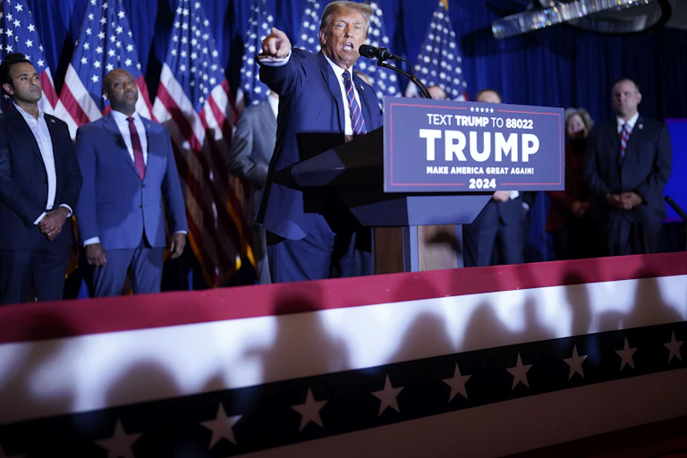 Donald Trump speaks at a lectern that says his name, standing in front of a row of people, including Vivek Ramaswamy and Tim Scott, and a row of American flags.