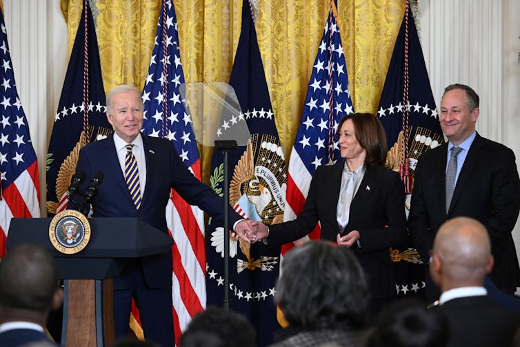 Joe Biden wears a dark suit and holds hands with Kamala Harris, also wearing a dark blazer, and standing next to her husband, Doug Emhoff. They stand in front of a podium and American flags.