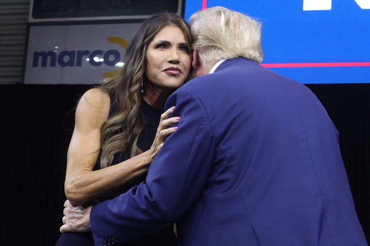 A white middle aged woman with dark hair wears a sleeveless dress and presses her face up against Donald Trump, seen only from the behind. He wears a navy blue jacket.