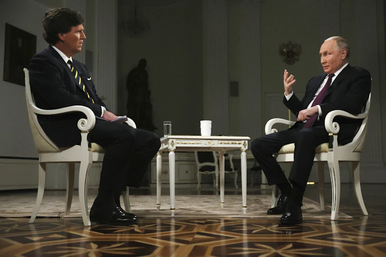Two men, one balding, sit in white chairs across from each other during an interview.