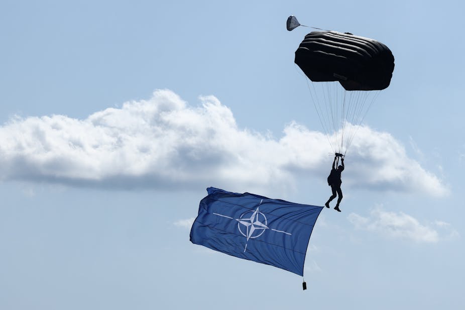 A large blue NATO flag is seen flying in the sky, below a person paratrooping.