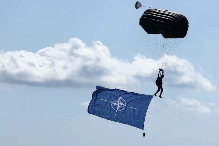 A large blue NATO flag is seen flying in the sky, below a person paratrooping.