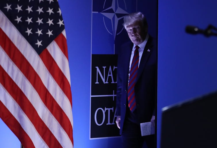 Donald Trump enters a stage with an American flag on it, with blue-lit lighting.