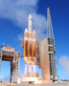 A gold and white rocket, with flames coming out of its end, attached to a concrete scaffold.