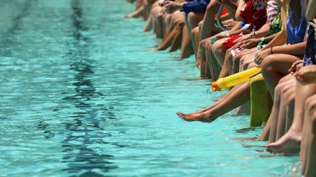 Row of people sitting at edge of swimming pool, legs in water