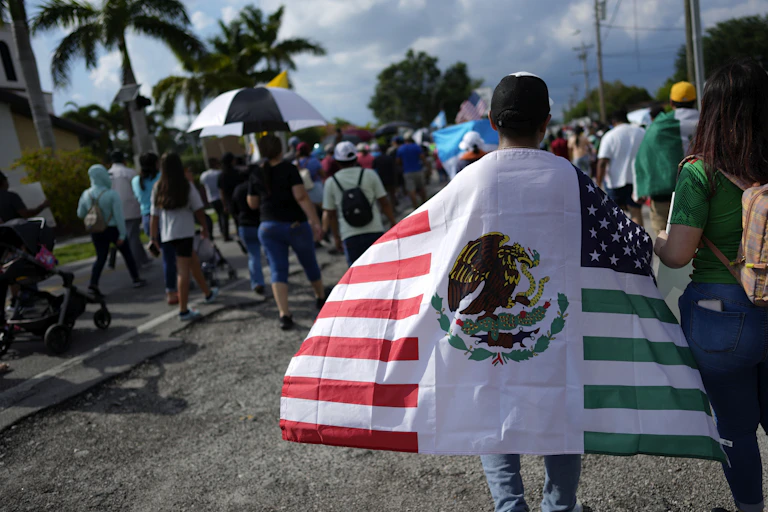 A man walks draped in a mashup of the United States and Mexico flags in a tropical place at the end of a big procession.