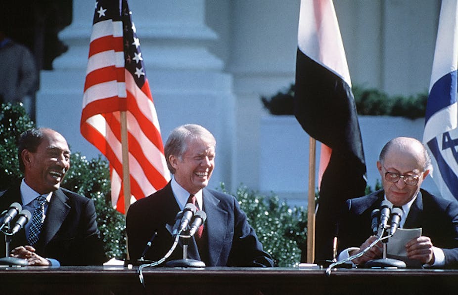 Three men in suits smiling in front of microphones; flags behind them
