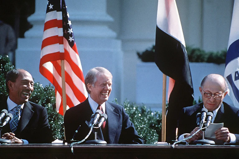 Three men in suits smiling in front of microphones; flags behind them