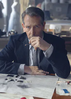 A gloomy looking man sits at a desk and has some fashion designs on the desk in front of him.