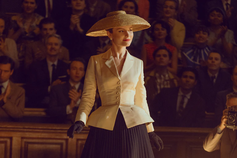A woman in a wide brimmed hat and a cream jacket with a cinched-in waist.