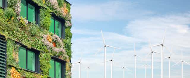 Left side, building with windows, walls covered in green plants, right side blue sky and a dozen white wind turbines