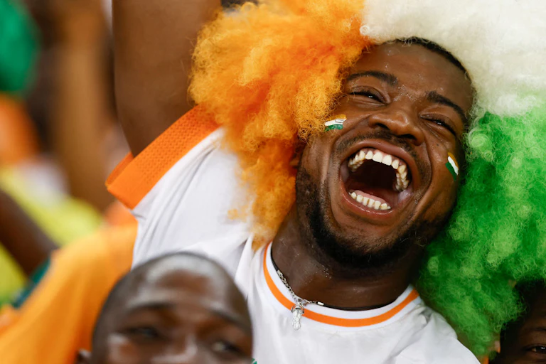 Un visage d'homme paré d'une perruque orange, blanche et verte. Sa bouche est ouverte sur un grand sourire, ses yeux sont plissés de joie.