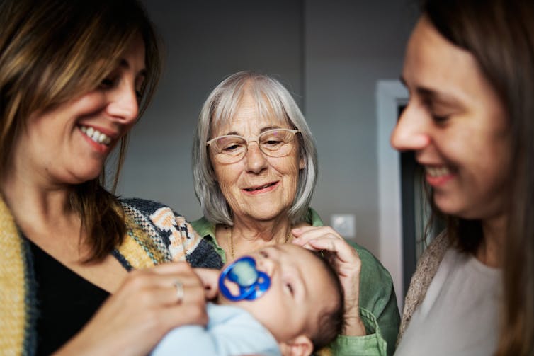 Three women, one of whom has white hair, stand smiling around a baby in a blue outfit and a pacifier.