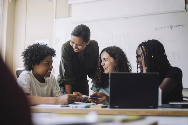 Three smiling students work at a laptop while their teacher stands behind them offering support.