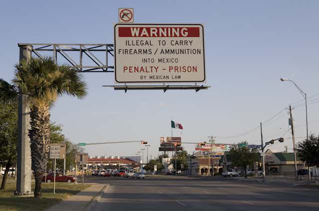 An English-language road sign warns drivers that it's illegal to bring guns from the U.S. to Mexico. A Mexican flag waves in the distance.