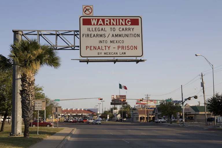 An English-language road sign warns drivers that it's illegal to bring guns from the U.S. to Mexico. A Mexican flag waves in the distance.
