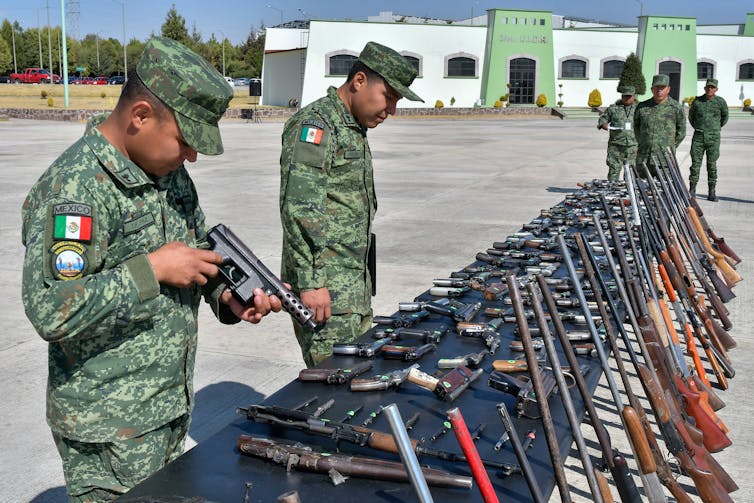 Two military agents wearing camouflage uniforms examine dozens of seized illegal guns displayed on a table.