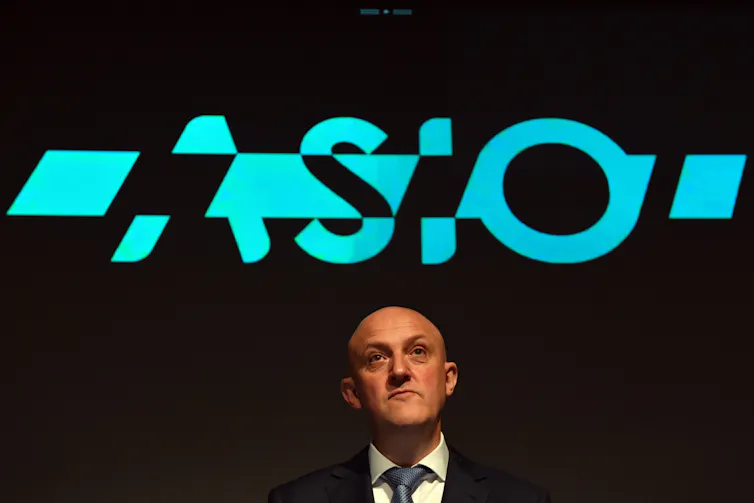 A photo of a bald middle-aged man in a suit in front of a sign reading ASIO.