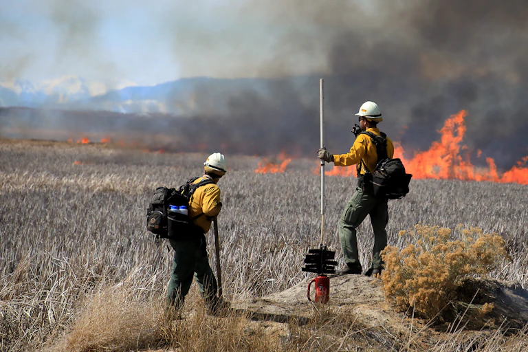 Two firefighters watch a line of flames through dry grass with mountains in the background.