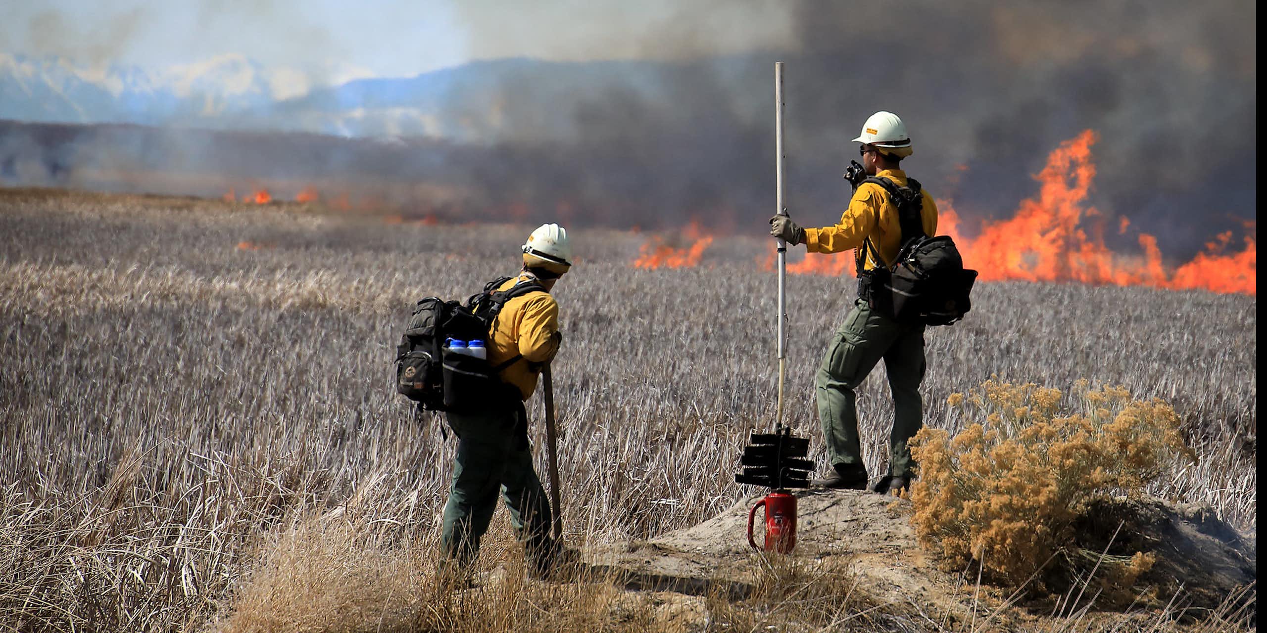 Two firefighters watch a line of flames through dry grass with mountains in the background.