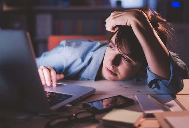 A tired woman looking at laptop late at night at a busy desk
