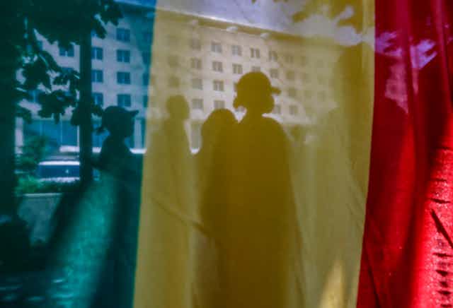Outlines of people standing in a street, blurred by a flag in the foreground.