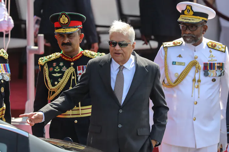 A Sri Lankan man dressed in a suit in front of two men dressed in military uniform.