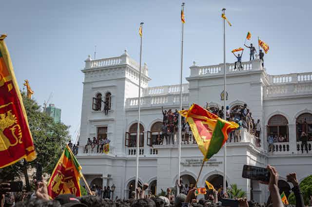 Protestors on the roof and hanging out of the windows of a large white building waving Sri Lanka flags.