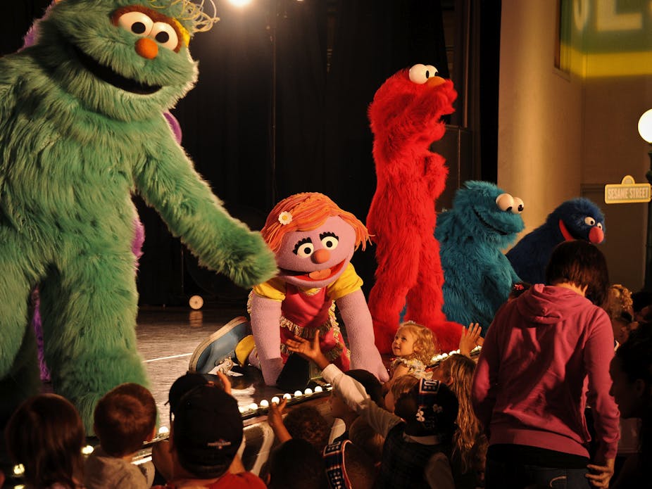 Large Sesame Street characters Rosita, Katie, Elmo, Cookie Monster and Grover stand on stage reaching out to children in the audience