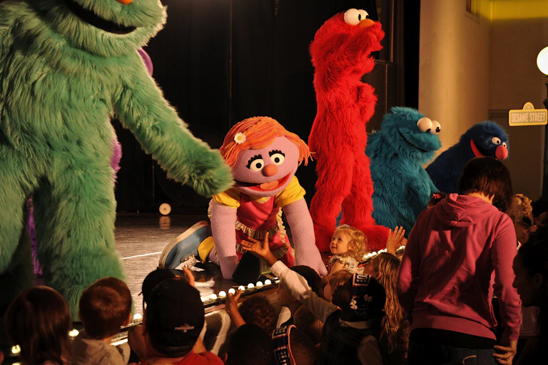 Large Sesame Street characters Rosita, Katie, Elmo, Cookie Monster and Grover stand on stage reaching out to children in the audience