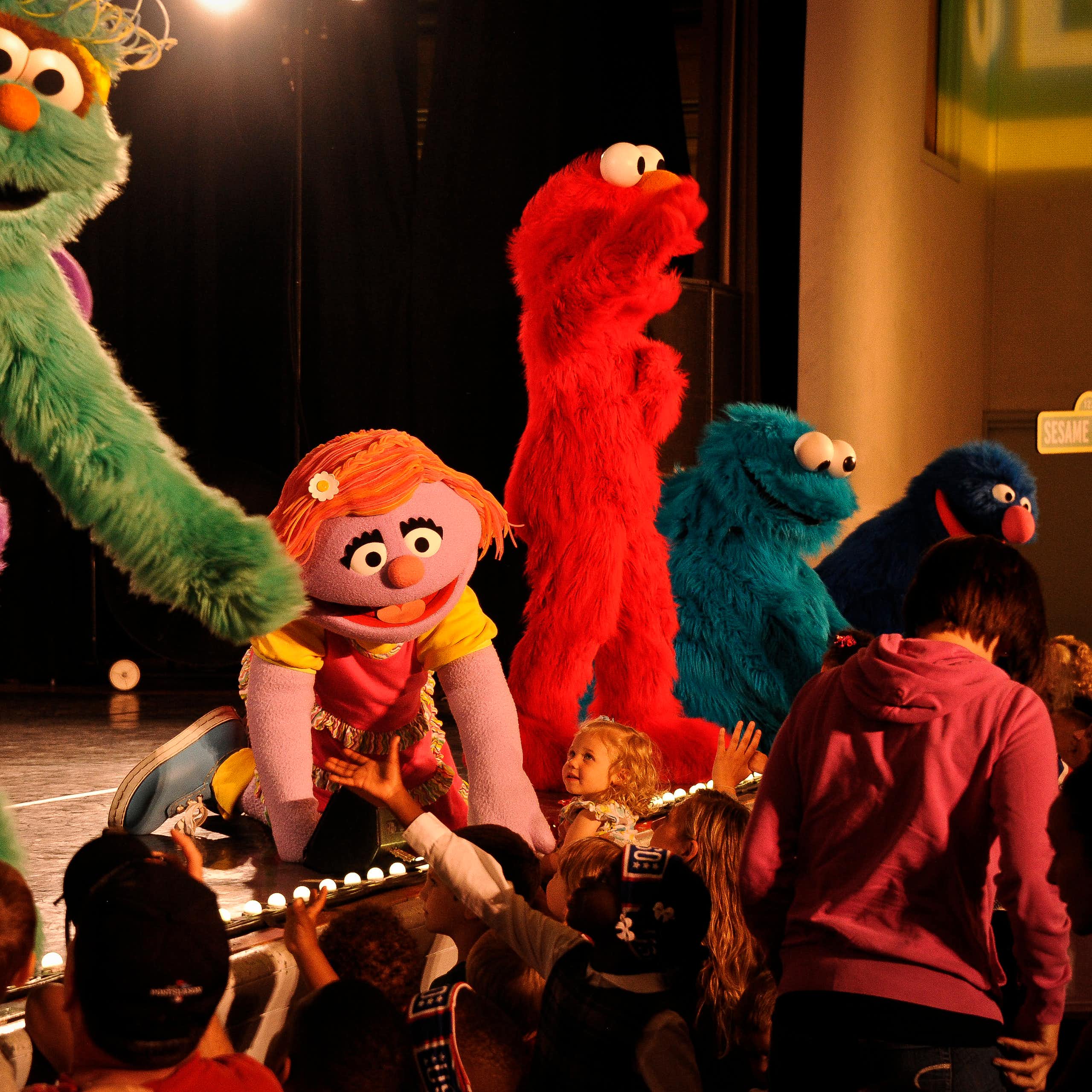 Large Sesame Street characters Rosita, Katie, Elmo, Cookie Monster and Grover stand on stage reaching out to children in the audience