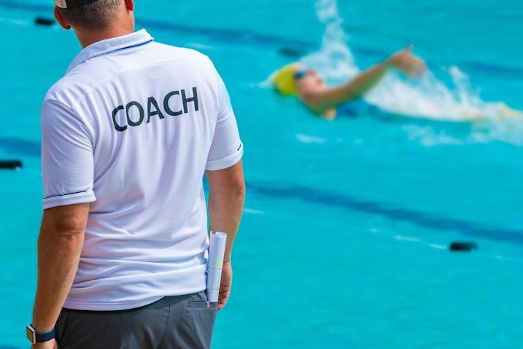A person in a t-shirt that says 'Coach' across the back faces toward a swimming pool and away from the camera